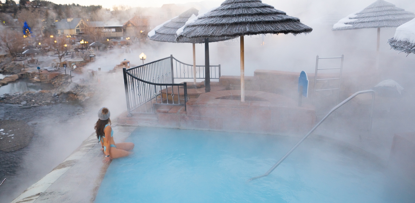 A rearview of a woman sitting and soaking her legs on the hot spring pool and watching the view