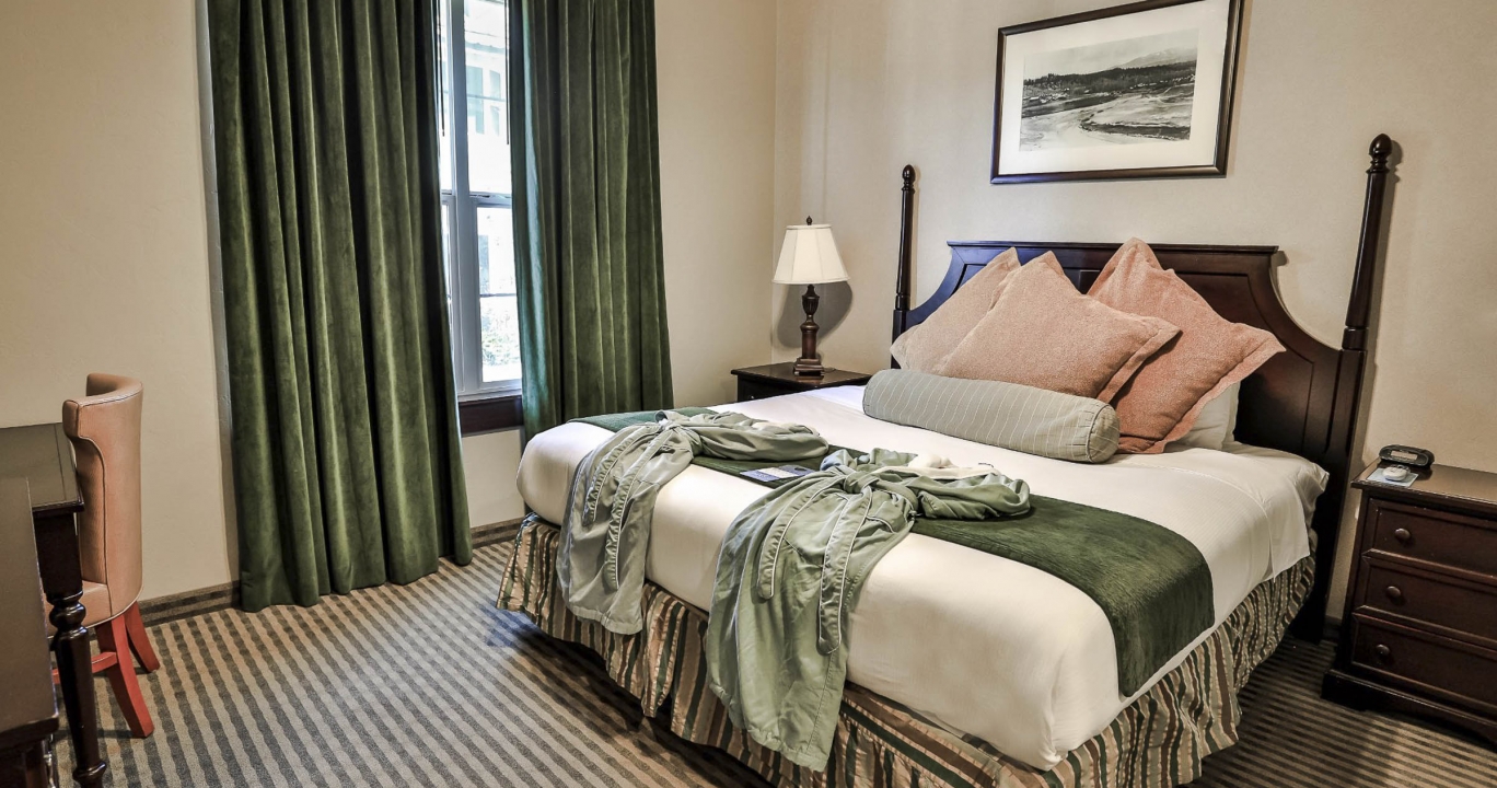 The bed between the two side tables with table lamps next to a large window with velvet curtains